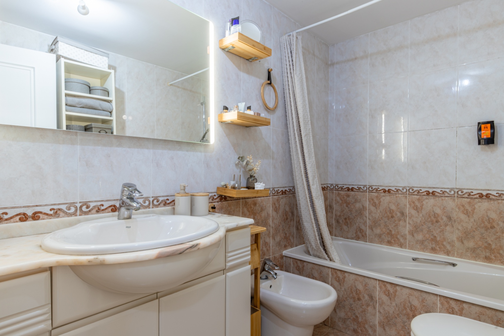 Complete badkamer met lichte tegels en decoratieve rand, geïntegreerde wastafel op marmeren blad, verlichte spiegel, ligbad met douchegordijn en bidet.