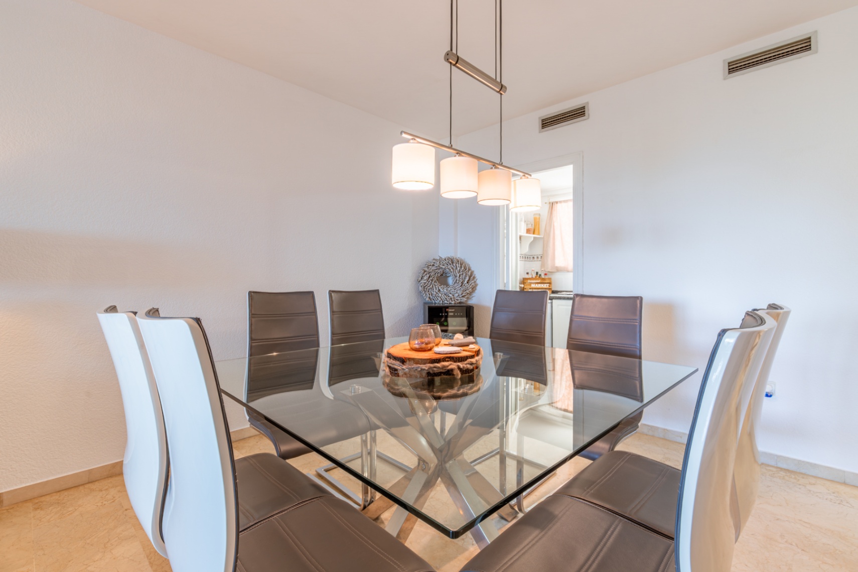 Eetkamer met vierkante glazen tafel en metalen onderstel, omringd door lederen stoelen. Een moderne hanglamp verlicht de ruimte, die aansluit op de keuken.