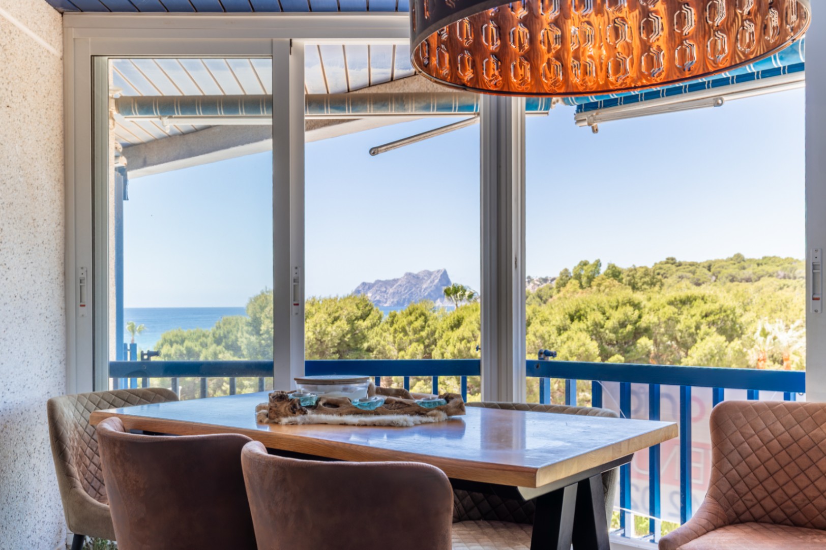 Eetruimte met panoramisch uitzicht op zee, bergen en weelderig groen via grote glazen schuifdeuren. Een unieke decoratieve hanglamp en comfortabele gestoffeerde stoelen sieren de ruimte.