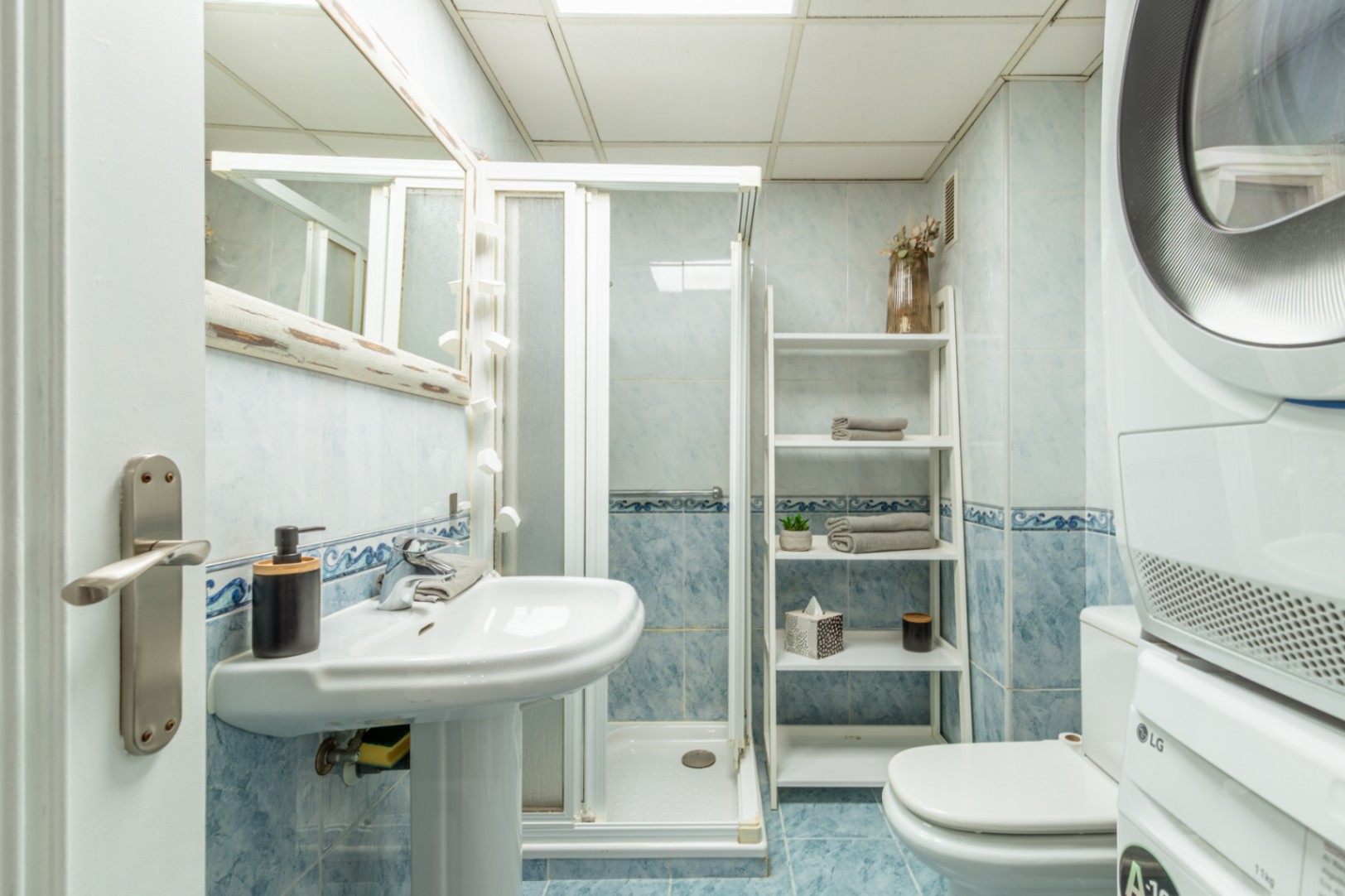 Functionele badkamer met lichtblauwe tegels, een voetstukwastafel, douchecabine en gestapelde LG-was- en droogapparaten. Een ladderrek biedt praktische opbergruimte.