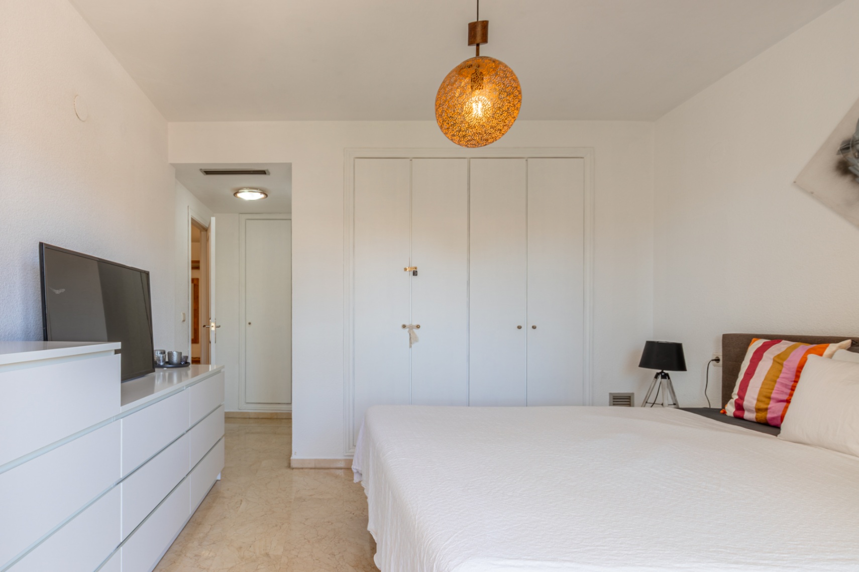 Hoofdslaapkamer met witte inbouwkasten, lichte marmeren vloer en een opvallende bolvormige hanglamp. Inclusief ladekast met tv en toegang tot gang.