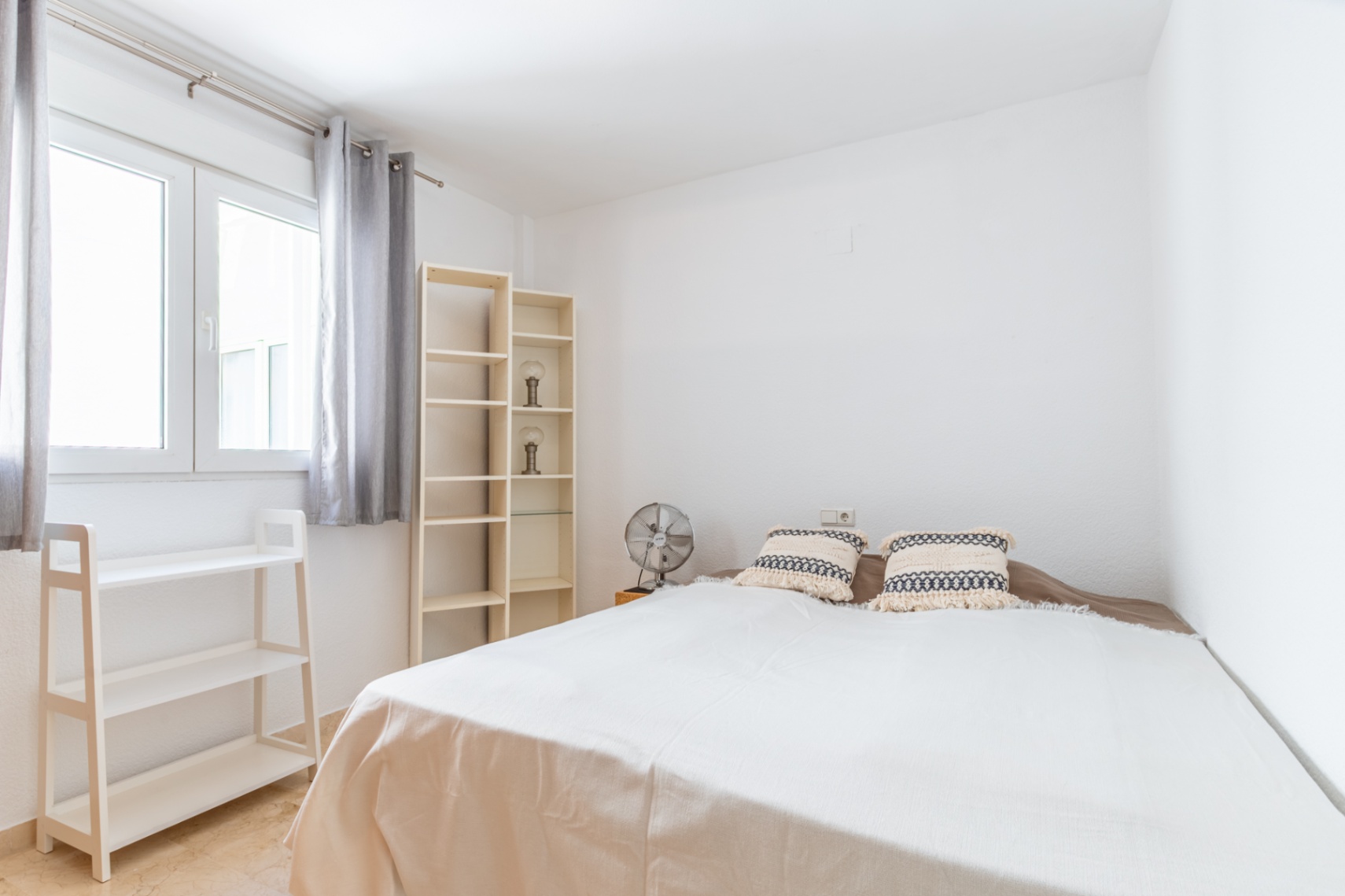 Lichte slaapkamer met tweepersoonsbed, lichte houten planken en ventilator. Het raam met grijze gordijnen biedt veel natuurlijk licht in de wit geschilderde ruimte met marmeren vloer.