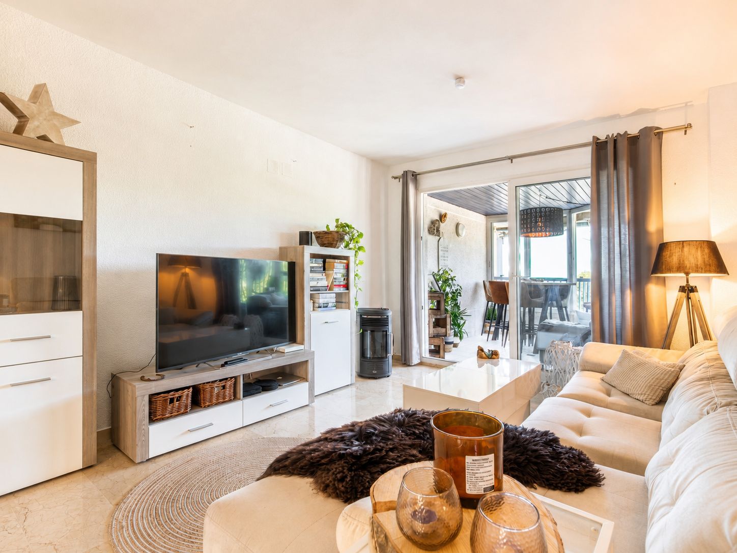 Lichte woonkamer met een lichte hoekbank, glanzende witte salontafel en tv-meubel. Glazen schuifdeuren bieden toegang tot een overdekt terras.