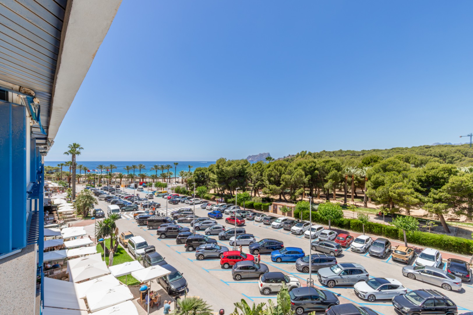 Panoramisch terrasuitzicht op de Middellandse Zee, een palmenstrand en de verre Peñón de Ifach. Beneden zijn een parkeerplaats en witte restaurantluifels zichtbaar.