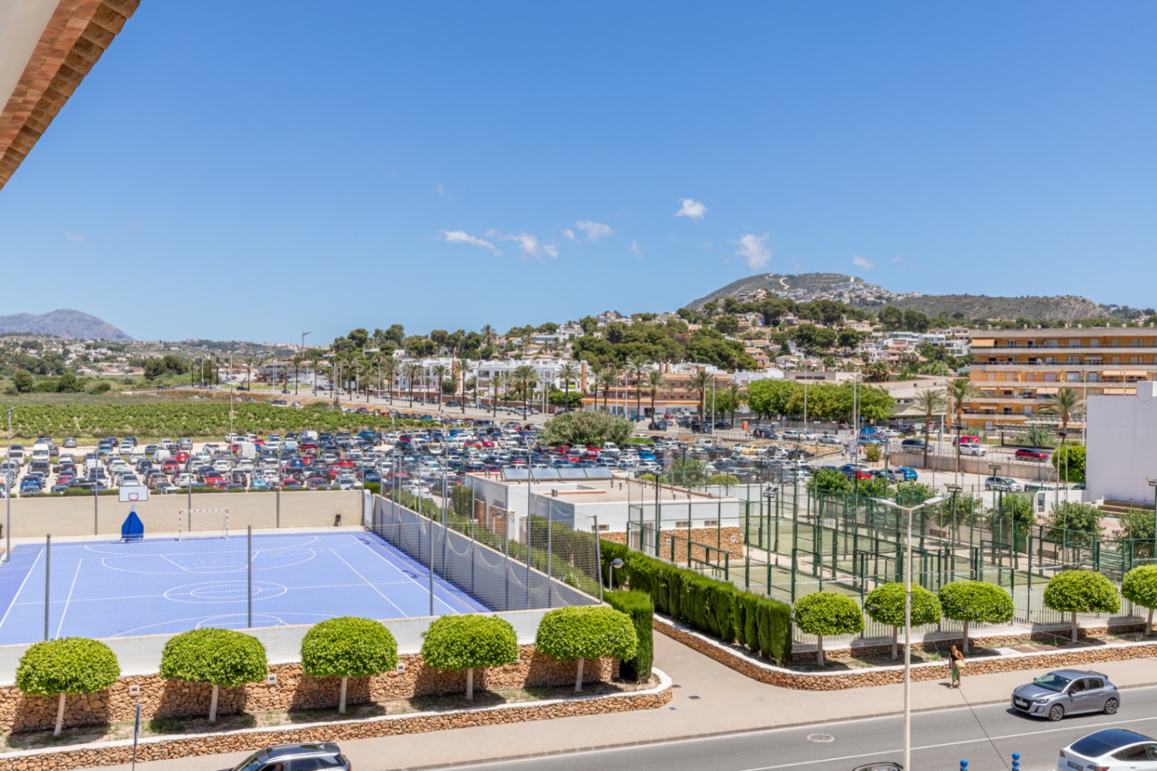 Panoramisch uitzicht met een blauw sportveld, meerdere padelbanen, een ruime parkeerplaats en het stadslandschap van Moraira met bergen op de achtergrond.