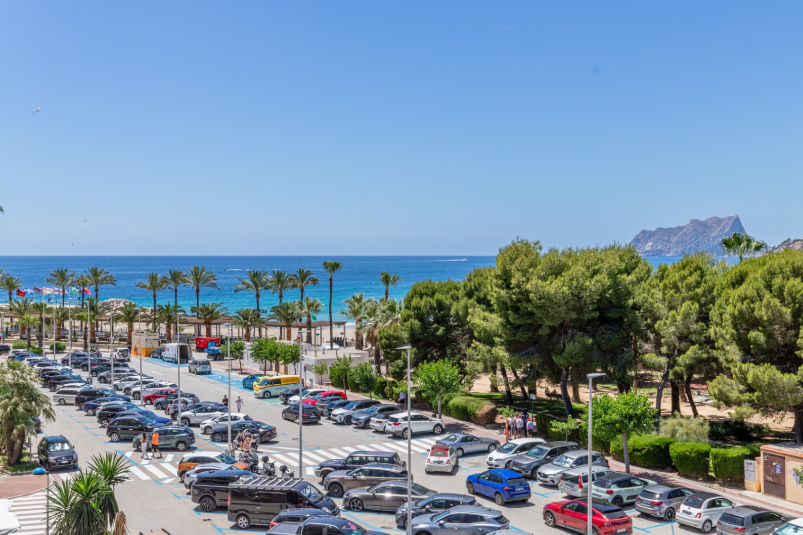 Panoramisch uitzicht op de Middellandse Zee, met palmbomen, zandstrand en de verre Peñón de Ifach, naast een drukke parkeerplaats en pijnbomen.