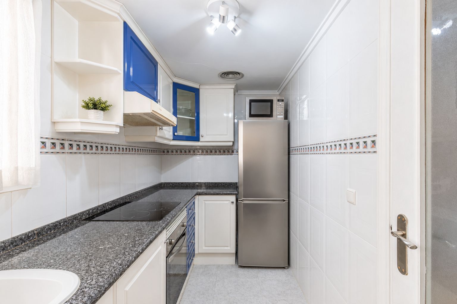 Praktische keuken met witte en blauwe kasten, donkere granieten werkbladen, een geïntegreerde inductiekookplaat en RVS-apparatuur. Natuurlijk licht valt binnen via het raam.
