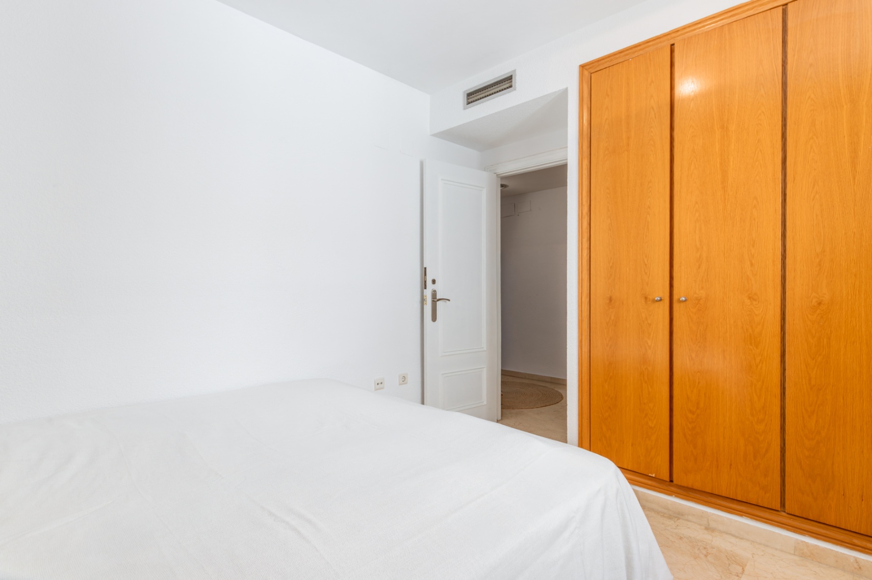 Slaapkamer met ruime, lichte houten inbouwkast en een bed met wit beddengoed. De kamer heeft een marmerlook vloer en een open deur naar een aangrenzende ruimte.
