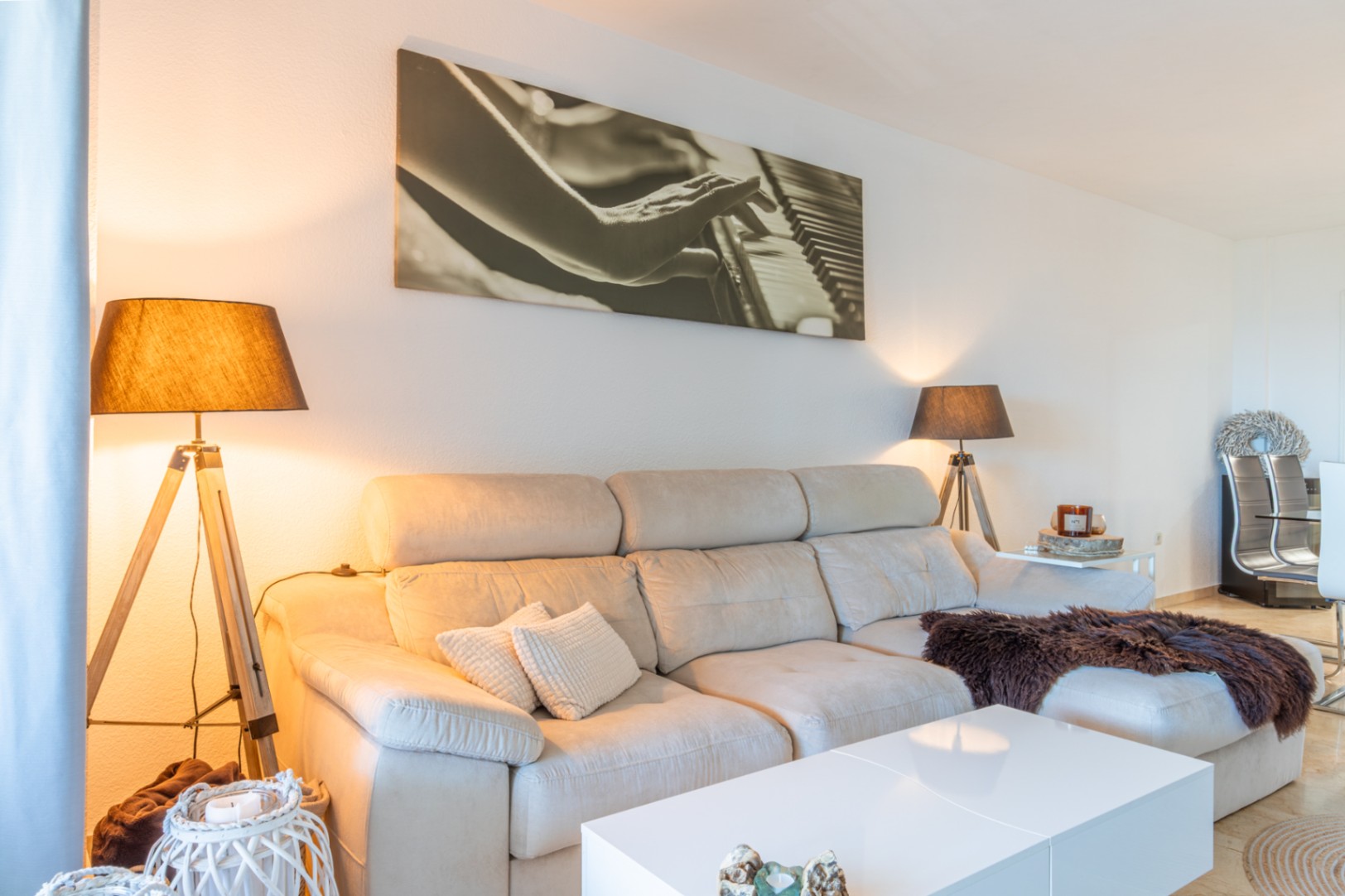 Woonkamer met lichte hoekbank, witte salontafel en twee vloerlampen met houten statief. Een groot kunstwerk van een piano siert de muur.