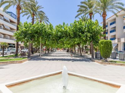 Brede voetgangerspromenade in Moraira, met een vierkante fontein op de voorgrond, beschaduwd door een bladerdak van groene bomen en hoge palmbomen, geflankeerd door gebouwen.
