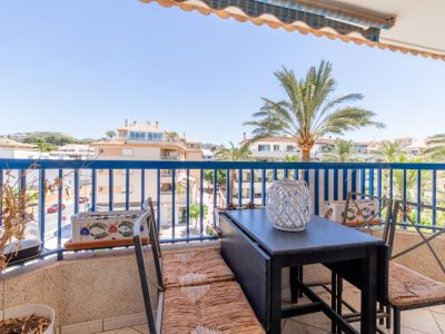 Buitenbalkon met opklapbare tafel, rieten stoelen, blauwe balustrade en uitzicht op de straat, gebouwen en palmbomen onder een luifel.