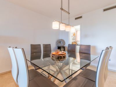 Eetkamer met vierkante glazen tafel en metalen onderstel, omringd door lederen stoelen. Een moderne hanglamp verlicht de ruimte, die aansluit op de keuken.