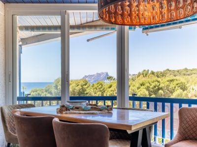 Eetruimte met panoramisch uitzicht op zee, bergen en weelderig groen via grote glazen schuifdeuren. Een unieke decoratieve hanglamp en comfortabele gestoffeerde stoelen sieren de ruimte.
