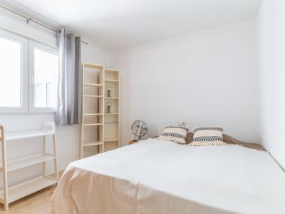 Lichte slaapkamer met tweepersoonsbed, lichte houten planken en ventilator. Het raam met grijze gordijnen biedt veel natuurlijk licht in de wit geschilderde ruimte met marmeren vloer.