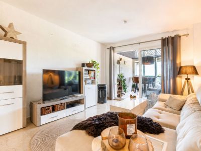 Lichte woonkamer met een lichte hoekbank, glanzende witte salontafel en tv-meubel. Glazen schuifdeuren bieden toegang tot een overdekt terras.