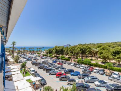 Panoramisch terrasuitzicht op de Middellandse Zee, een palmenstrand en de verre Peñón de Ifach. Beneden zijn een parkeerplaats en witte restaurantluifels zichtbaar.