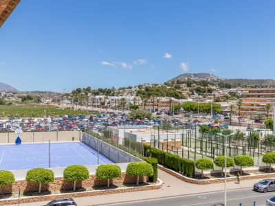 Panoramisch uitzicht met een blauw sportveld, meerdere padelbanen, een ruime parkeerplaats en het stadslandschap van Moraira met bergen op de achtergrond.