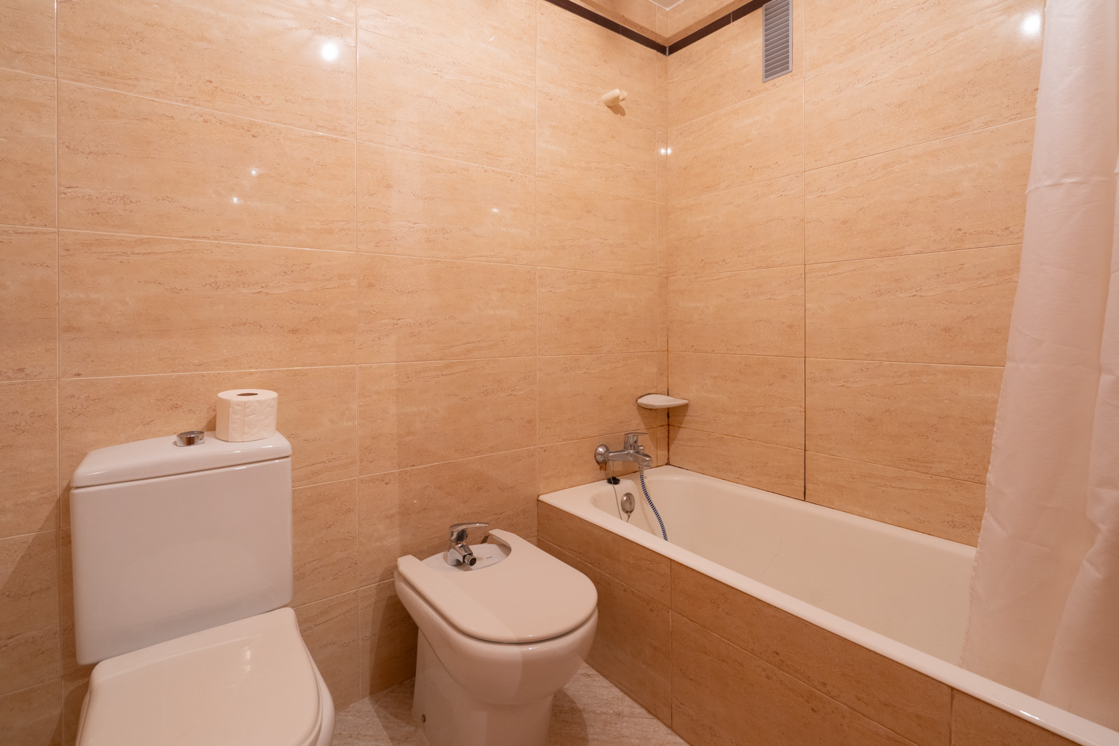 Badkamer met beige betegelde muren, voorzien van toilet, bidet en ligbad met douchegordijn. Verchroomde kranen en een hoekplankje zijn zichtbaar.