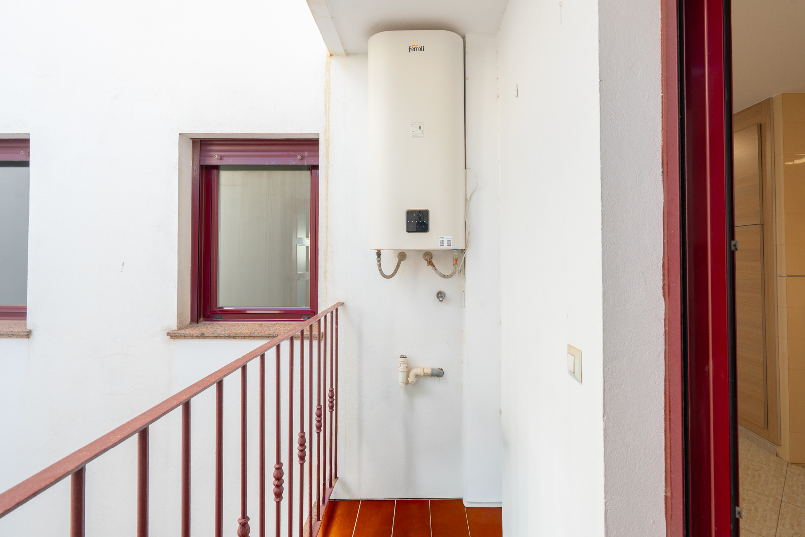 Binnenplaats met een aan de muur gemonteerde Ferroli boiler, terracotta vloertegels en een rood metalen balustrade. Een bordeauxrood raam met granieten vensterbank en een bijpassend deurkozijn zijn zichtbaar.
