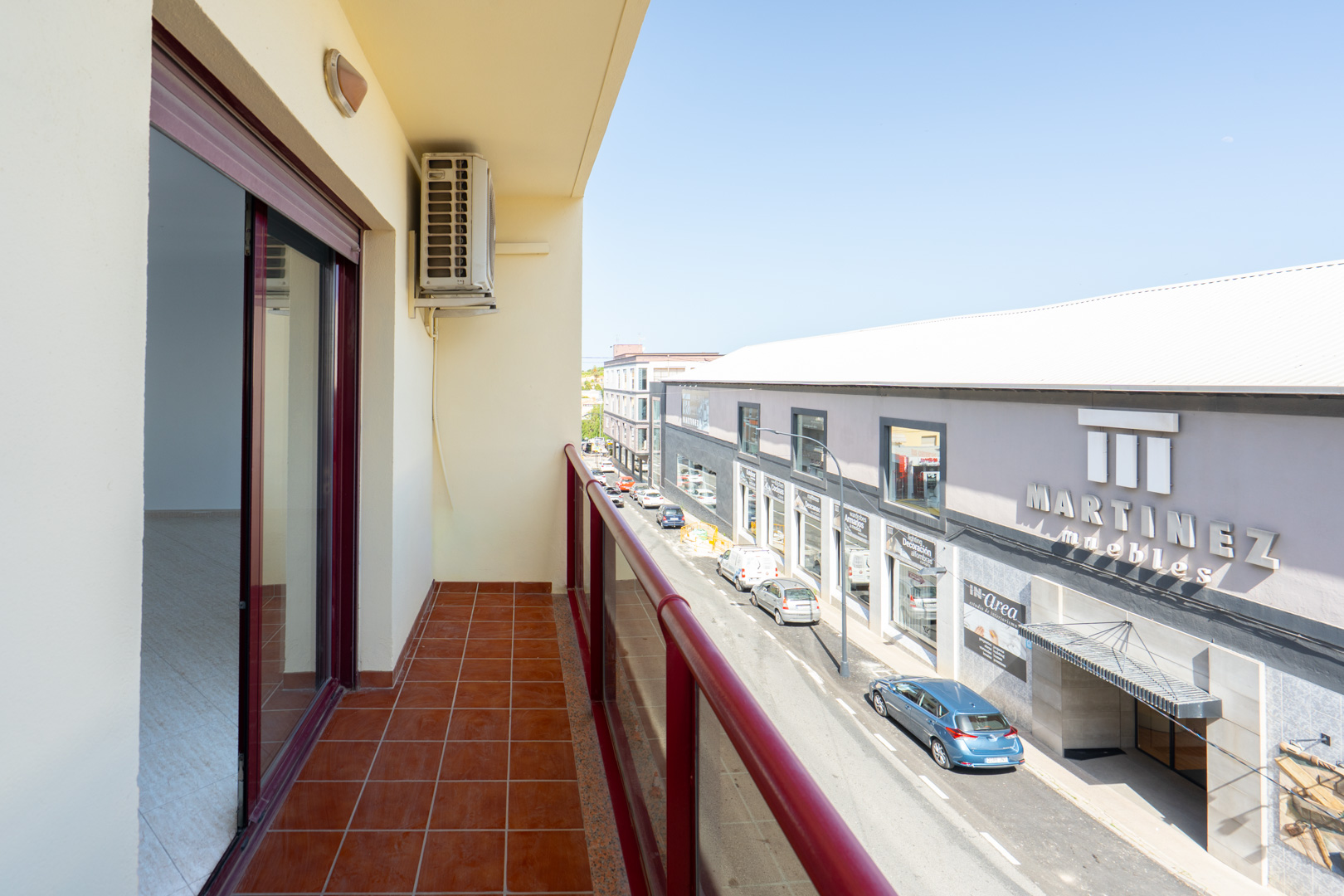 Buitenbalkon met terracottategels en rode balustrade. Een airconditioningunit is zichtbaar, met uitzicht op de straat en de winkel "Martinez Muebles".