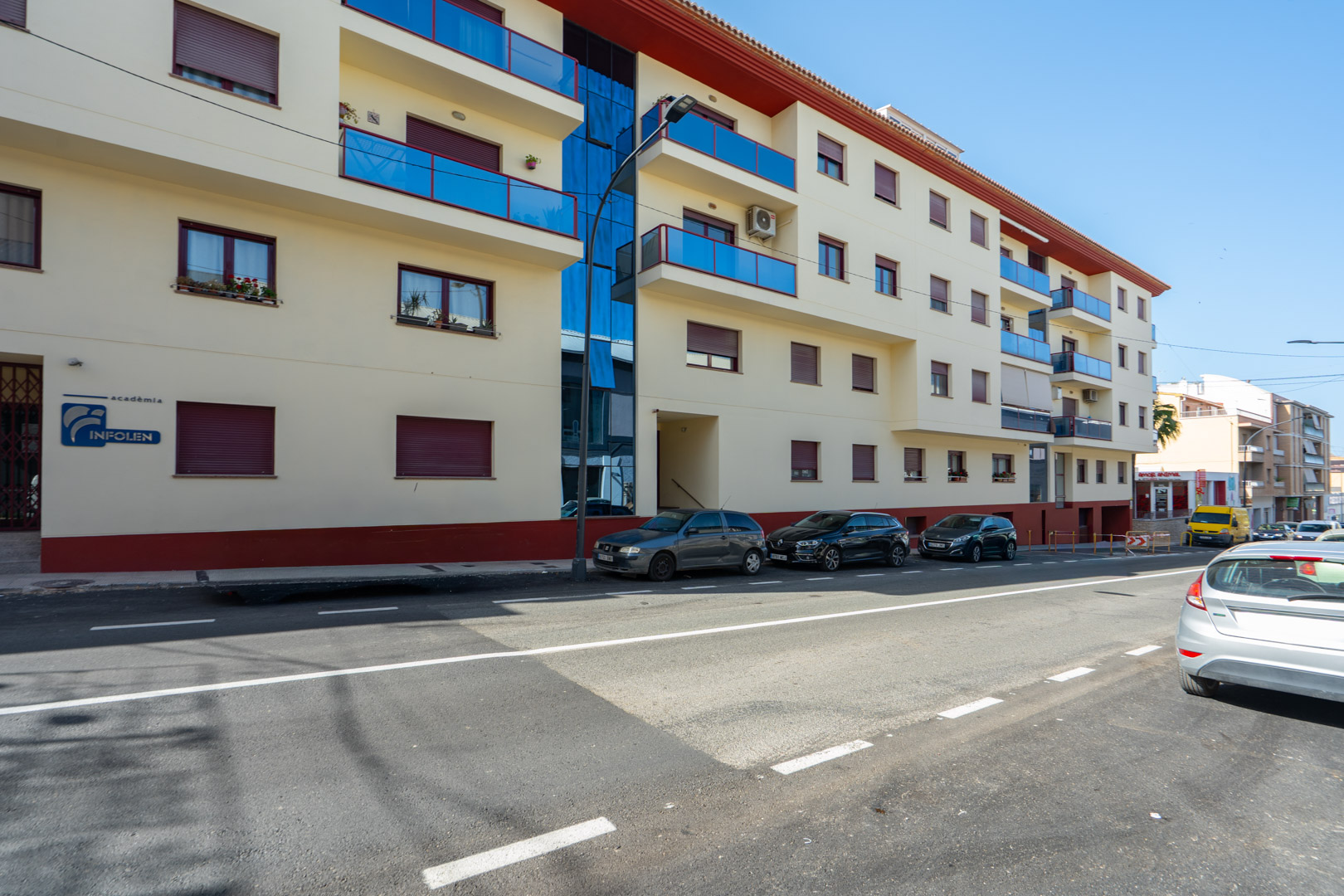 Exterieur van het woongebouw met lichte gevel, blauwe glazen balkons, ramen met bordeauxrode rolluiken en een verticaal glazen paneel.