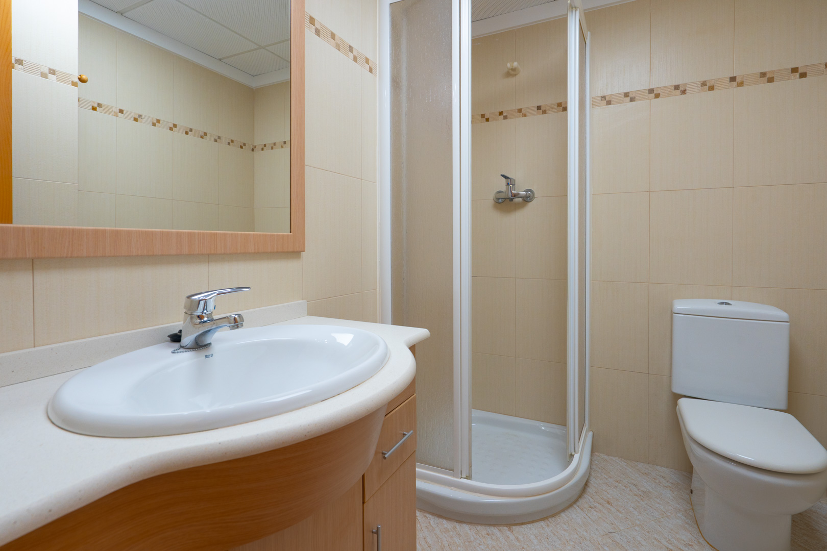 Functionele badkamer met ovale wastafel op licht houten meubel, ingelijste spiegel, hoekdouche en beige tegels met decoratieve rand.