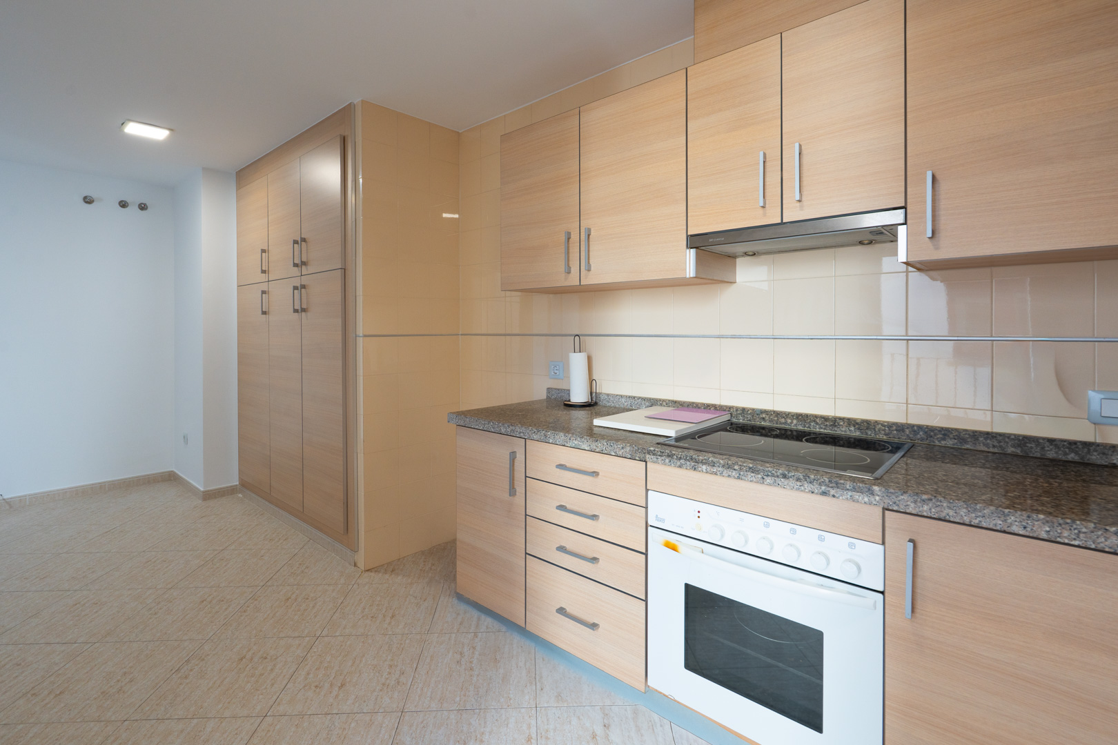 Functionele keuken met licht houtlook kasten, graniet-effect aanrechtblad, geïntegreerde keramische kookplaat en oven. Hoge opbergkast en beige vloertegels.
