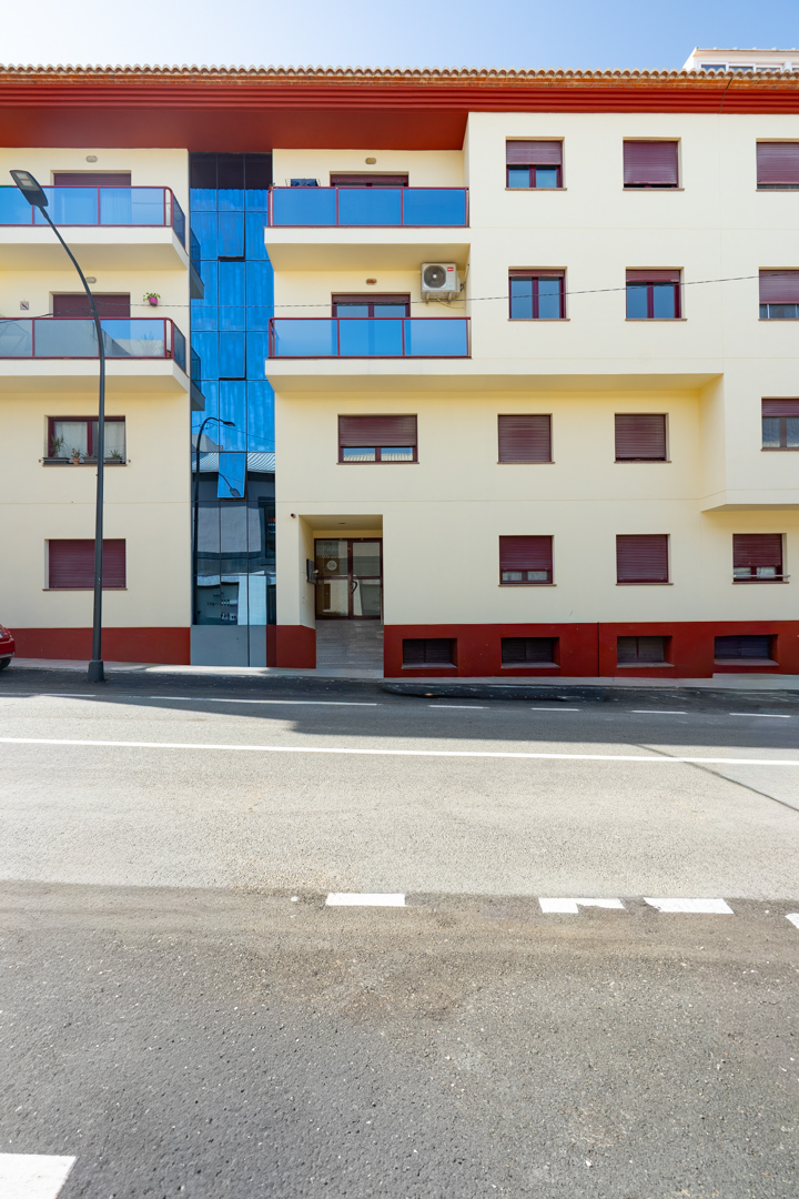 Gevel van het woongebouw met crèmekleurige afwerking, blauwe glazen balkons en rode rolluiken. Een centraal blauw glazen gedeelte en een entree op straatniveau met oprit zijn zichtbaar.
