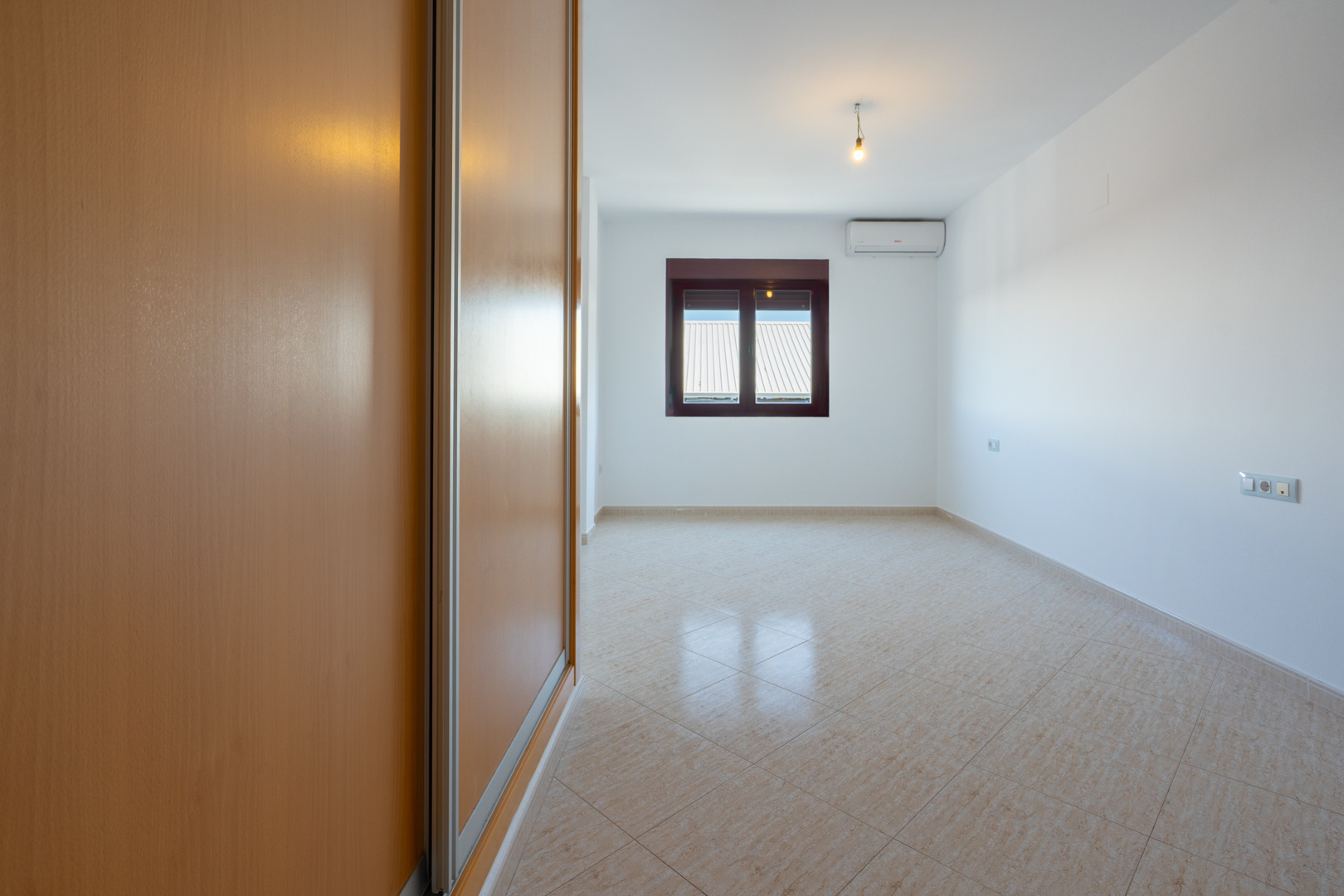 Lege kamer met lichte houten inbouwkast, diagonaal gelegde beige tegels en raam met uitzicht op daken. Een airconditioning unit is aanwezig.