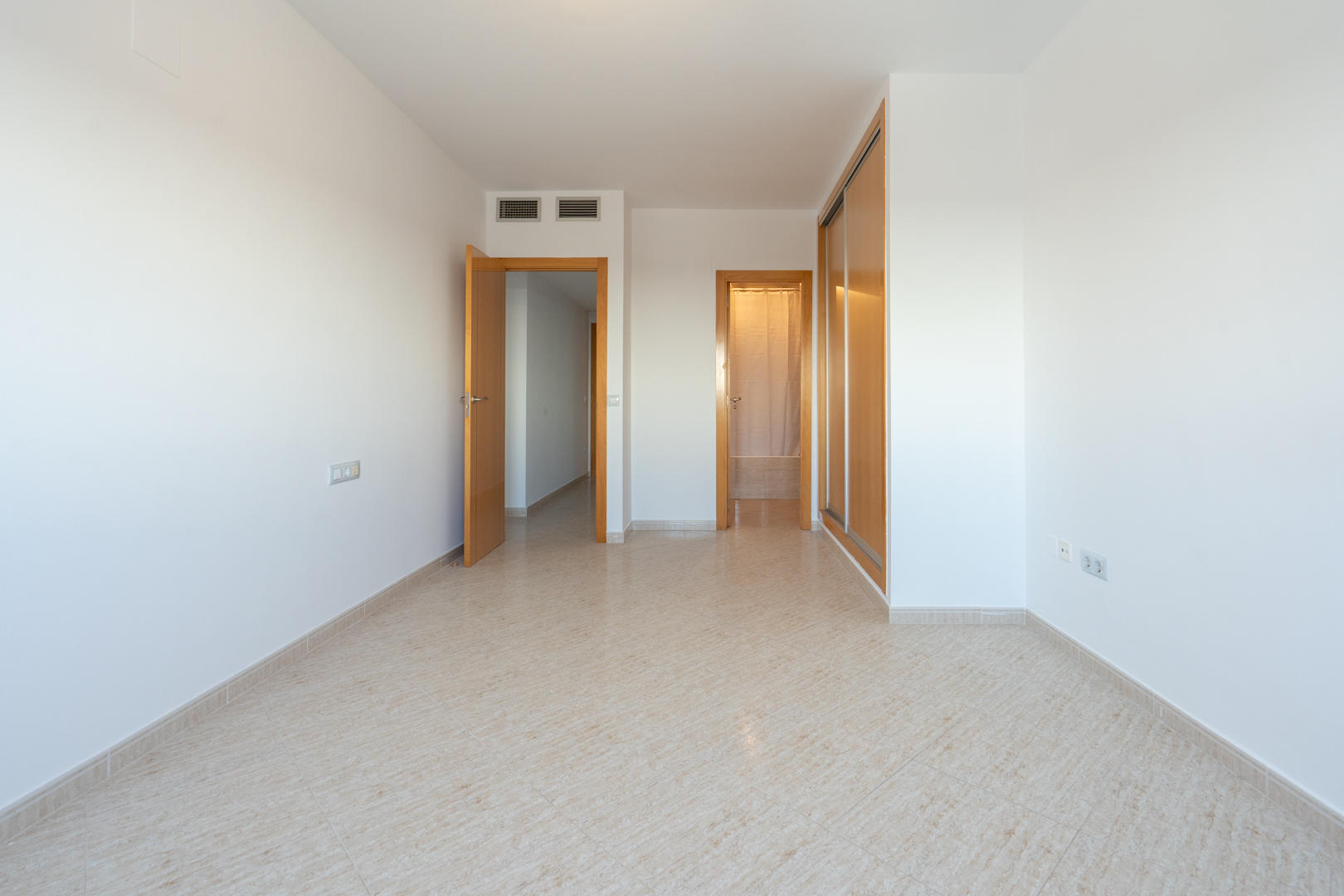Lege kamer met lichte tegelvloer, een ingebouwde houten kledingkast met schuifdeuren en deuren naar aangrenzende ruimtes. Airconditioningroosters zijn zichtbaar.