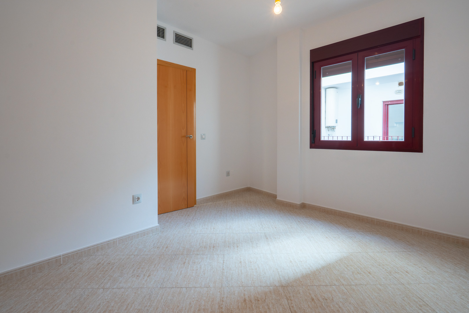 Lege kamer met lichte tegelvloer en witte muren. Voorzien van een houten deur en een raam met bordeauxrode kozijnen, uitkijkend op een binnenplaats met boiler. Ventilatieopeningen zichtbaar.