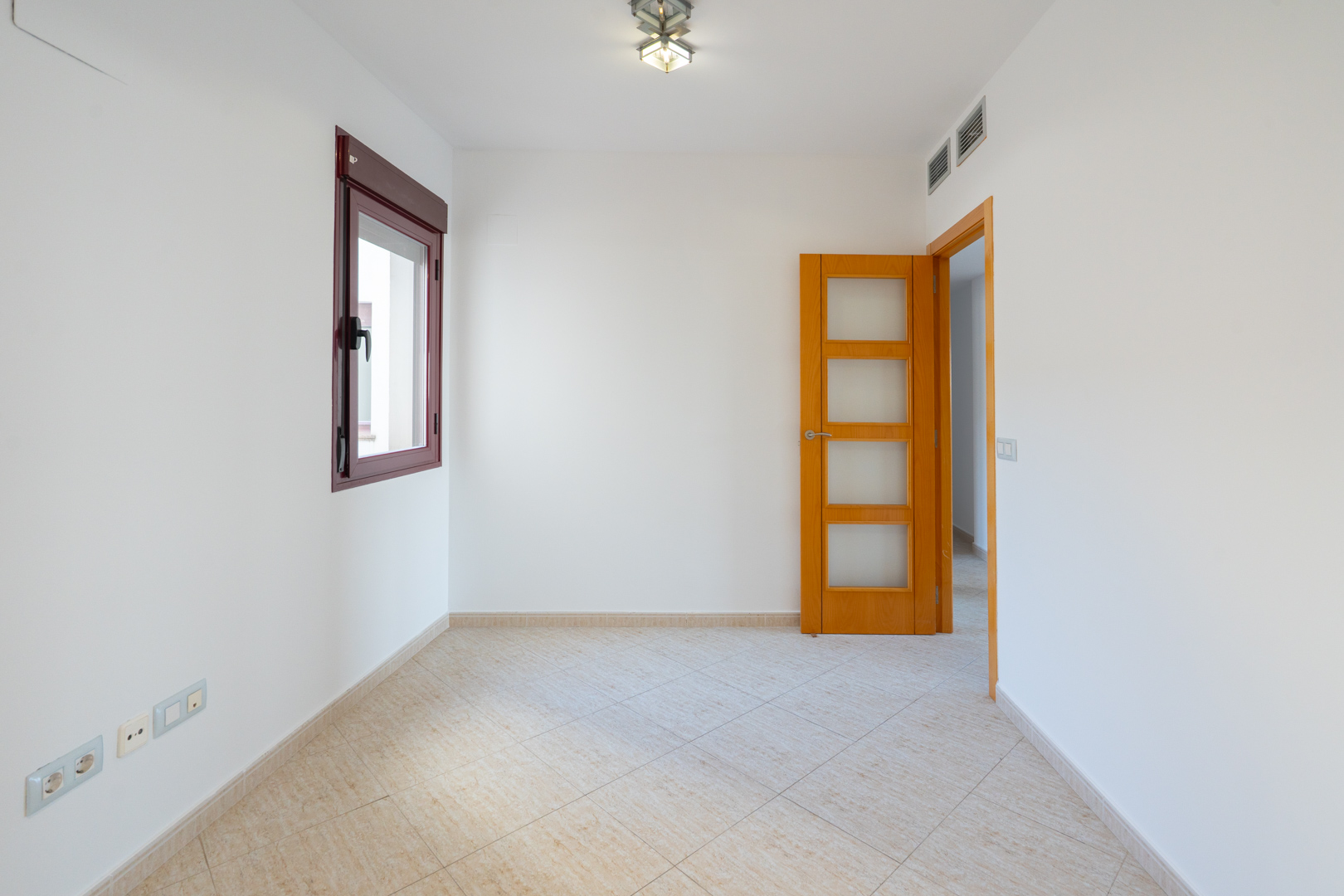 Lege kamer met witte muren, bordeauxrood kozijn en lichtbeige diagonale vloertegels. Een houten deur met glazen panelen en airco-roosters zijn aanwezig.