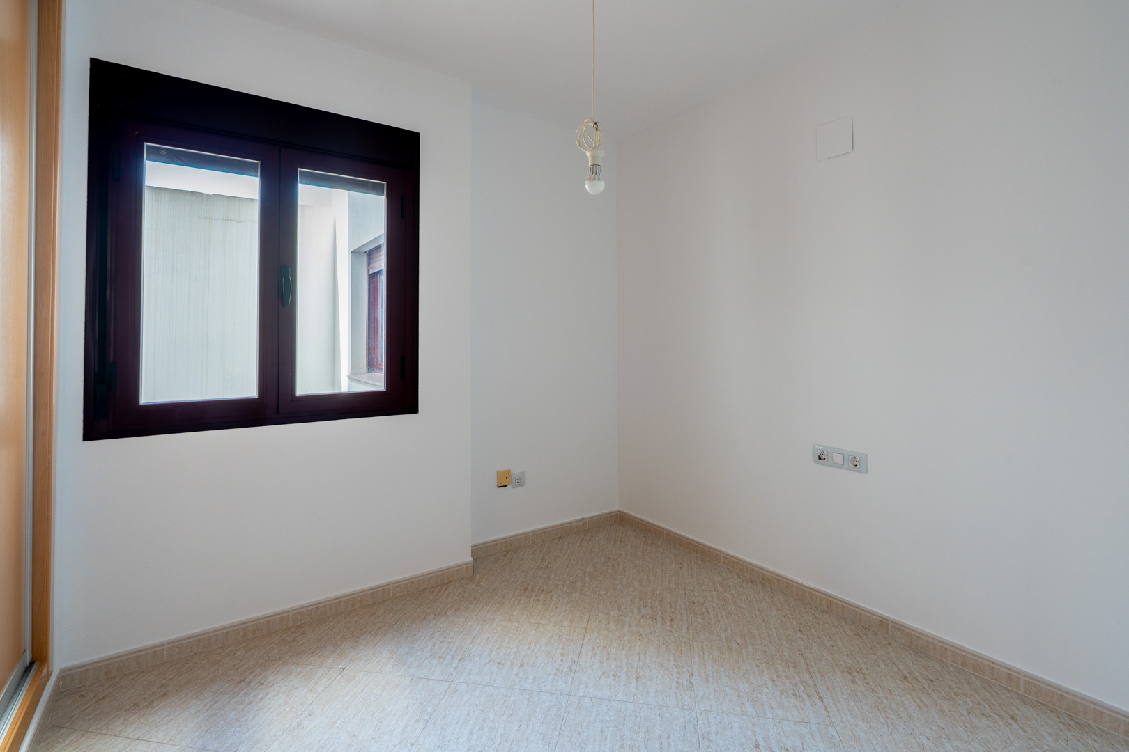 Lege kamer met witte muren en lichte tegelvloer. Het raam met donker kozijn kijkt uit op een binnenplaats. Een enkele lamp hangt aan het plafond.