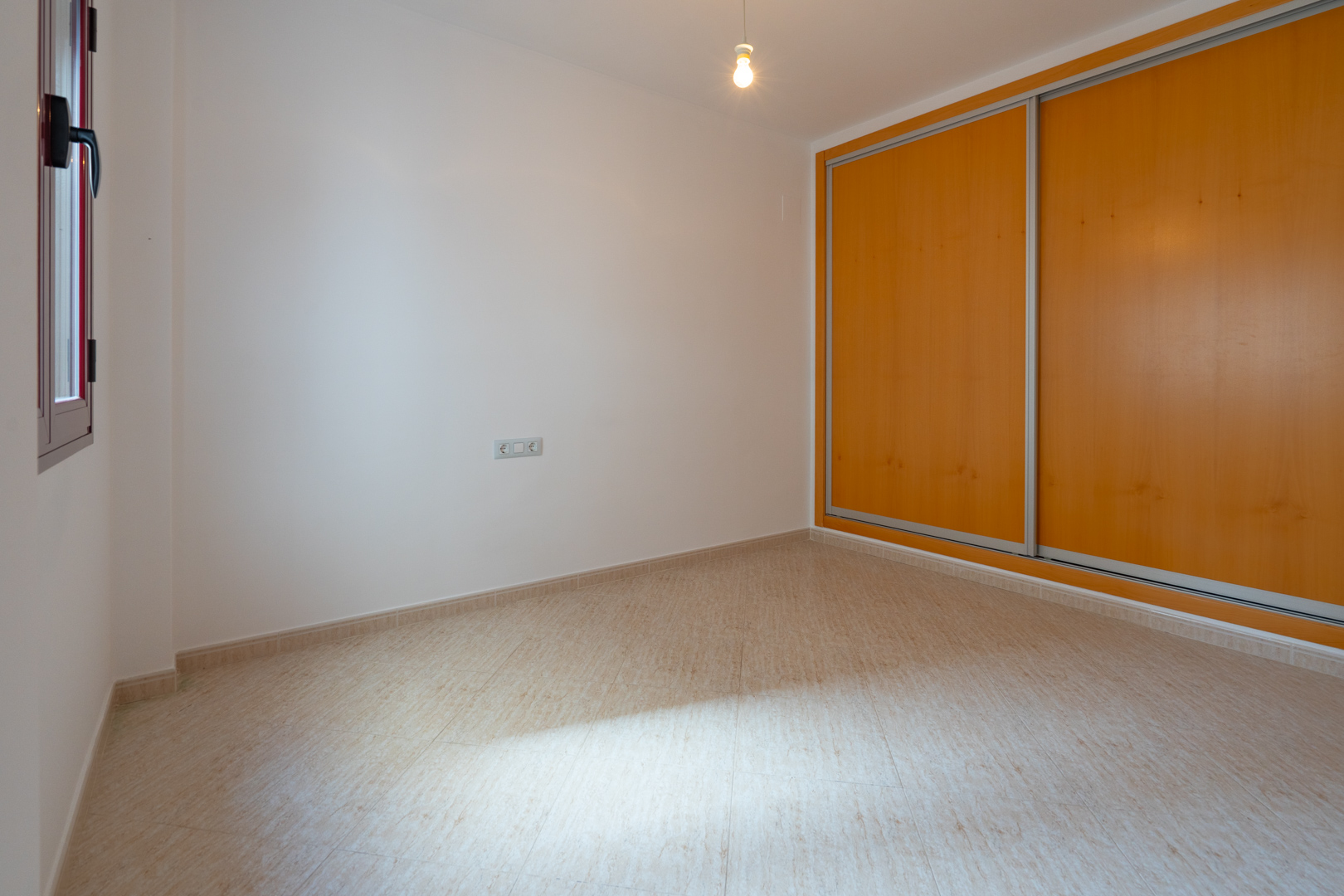 Lichte kamer met een ingebouwde kledingkast voorzien van twee lichte houten schuifdeuren en een beige tegelvloer. Een raam met donker kozijn zorgt voor natuurlijke lichtinval.