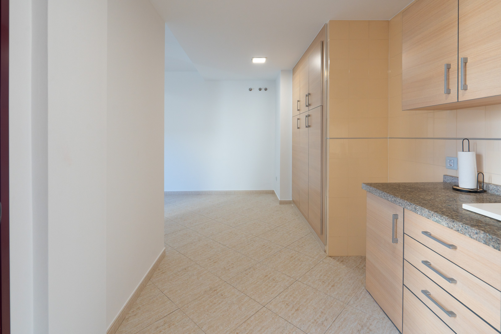 Overgang van de keuken met granieten werkblad en lichte houten kasten naar een hal met beige keramische vloer en wandhaken.