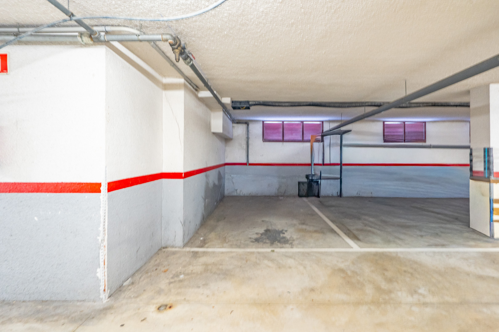 Privé ondergrondse parkeerplaats met betonnen vloer, wit-grijze muren met rode streep, en zichtbare leidingen aan het plafond. Twee kleine ramen met rode jaloezieën zijn zichtbaar.
