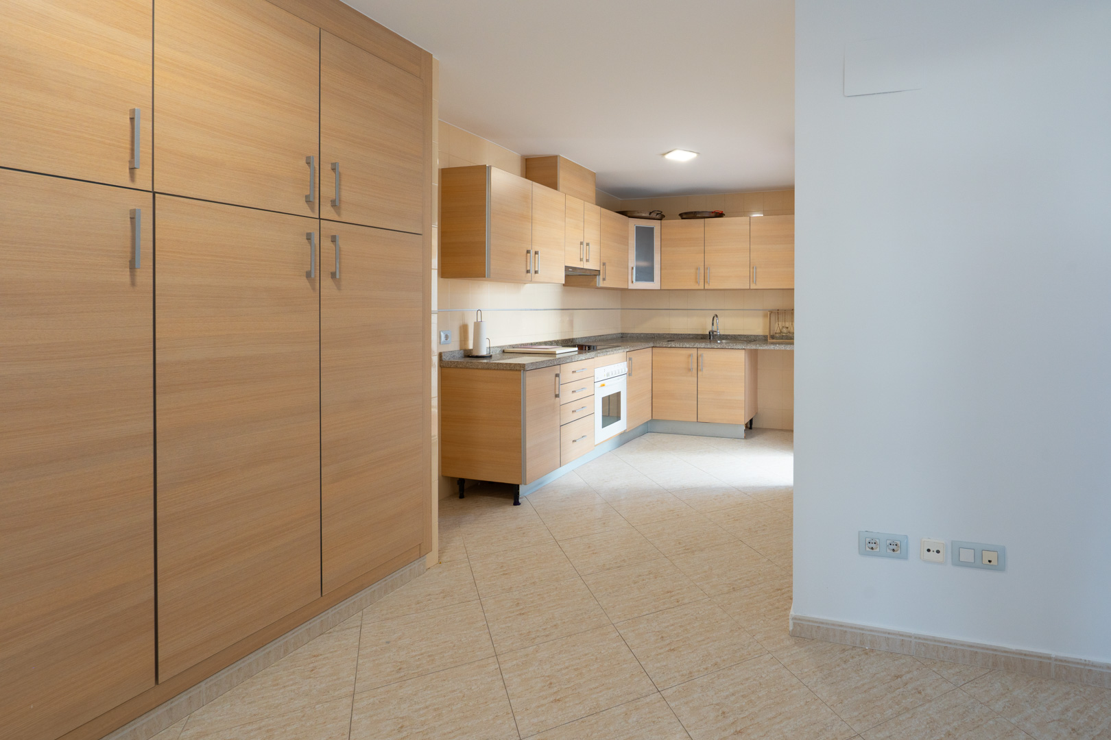 Ruime keuken met lichte houten kasten, gespikkelde granieten werkbladen en een geïntegreerde oven. Een grote bijpassende voorraadkast staat links op de beige tegelvloer.