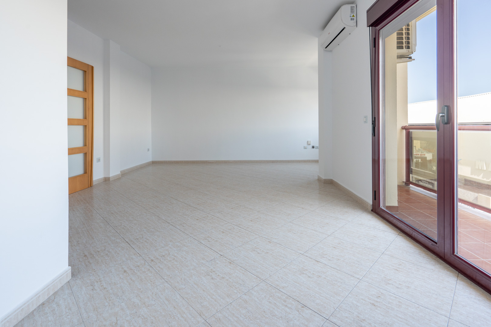 Ruime, ongemeubileerde woon-/eetkamer met lichte tegelvloer, houten deur met matglas panelen en toegang tot terras via donkerrode aluminium schuifdeur. Een airco-unit is zichtbaar.