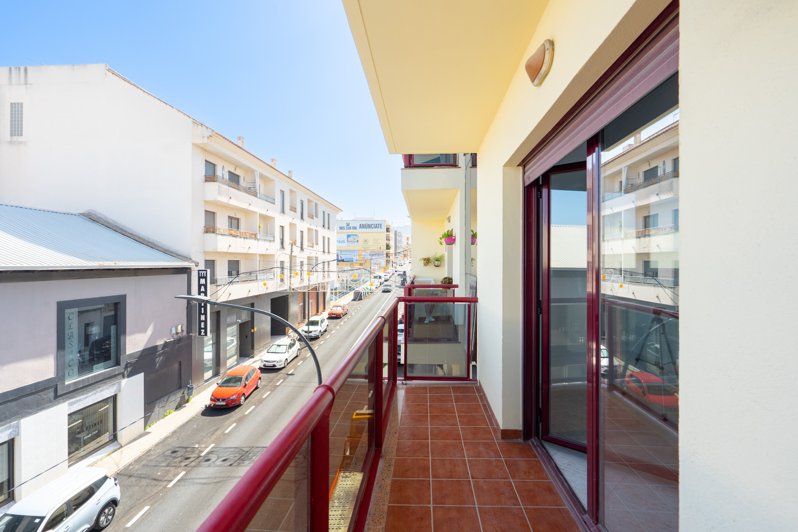 Zonnig balkon met terracotta vloertegels en een rood omrand glazen balustrade, met uitzicht op een stedelijke straat met gebouwen.