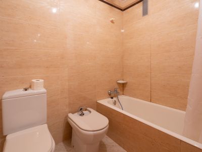 Badkamer met beige betegelde muren, voorzien van toilet, bidet en ligbad met douchegordijn. Verchroomde kranen en een hoekplankje zijn zichtbaar.