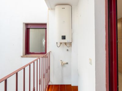 Binnenplaats met een aan de muur gemonteerde Ferroli boiler, terracotta vloertegels en een rood metalen balustrade. Een bordeauxrood raam met granieten vensterbank en een bijpassend deurkozijn zijn zichtbaar.