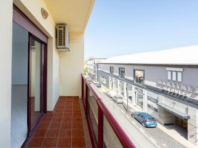 Buitenbalkon met terracottategels en rode balustrade. Een airconditioningunit is zichtbaar, met uitzicht op de straat en de winkel "Martinez Muebles".