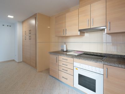 Functionele keuken met licht houtlook kasten, graniet-effect aanrechtblad, geïntegreerde keramische kookplaat en oven. Hoge opbergkast en beige vloertegels.