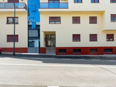 Gevel van het woongebouw met crèmekleurige afwerking, blauwe glazen balkons en rode rolluiken. Een centraal blauw glazen gedeelte en een entree op straatniveau met oprit zijn zichtbaar.