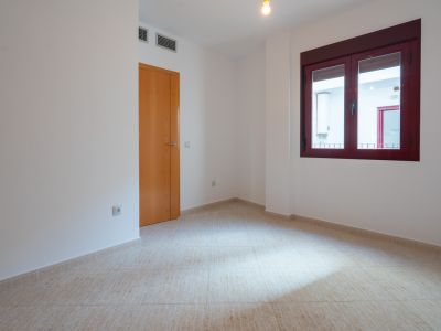 Lege kamer met lichte tegelvloer en witte muren. Voorzien van een houten deur en een raam met bordeauxrode kozijnen, uitkijkend op een binnenplaats met boiler. Ventilatieopeningen zichtbaar.