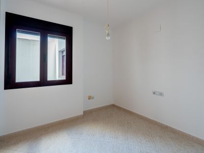 Lege kamer met witte muren en lichte tegelvloer. Het raam met donker kozijn kijkt uit op een binnenplaats. Een enkele lamp hangt aan het plafond.