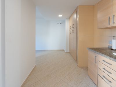 Overgang van de keuken met granieten werkblad en lichte houten kasten naar een hal met beige keramische vloer en wandhaken.