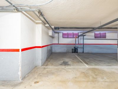 Privé ondergrondse parkeerplaats met betonnen vloer, wit-grijze muren met rode streep, en zichtbare leidingen aan het plafond. Twee kleine ramen met rode jaloezieën zijn zichtbaar.