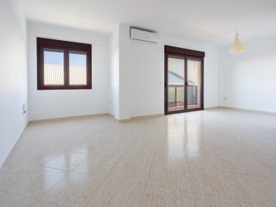 Ruime kamer met lichte tegelvloer, witte muren, een raam en een schuifdeur met bruine kozijnen naar een terras. Een wandgemonteerde airconditioning is aanwezig.