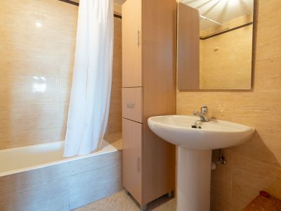 Tweede badkamer met wit voetstukwastafel, hoge houten opbergkast en ligbad met douchegordijn. De muren zijn afgewerkt met lichtbeige tegels.